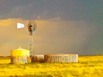 Windmill and silo with words "Water is Life" graffitied with a storm in the distance.